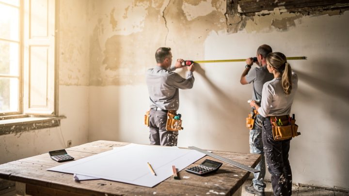 Maison ancienne en rénovation, avec trois artisans mesurant les pièces près d’une table en bois couverte de plans vierges et d’un calculateur, éclairée par la lumière naturelle.
